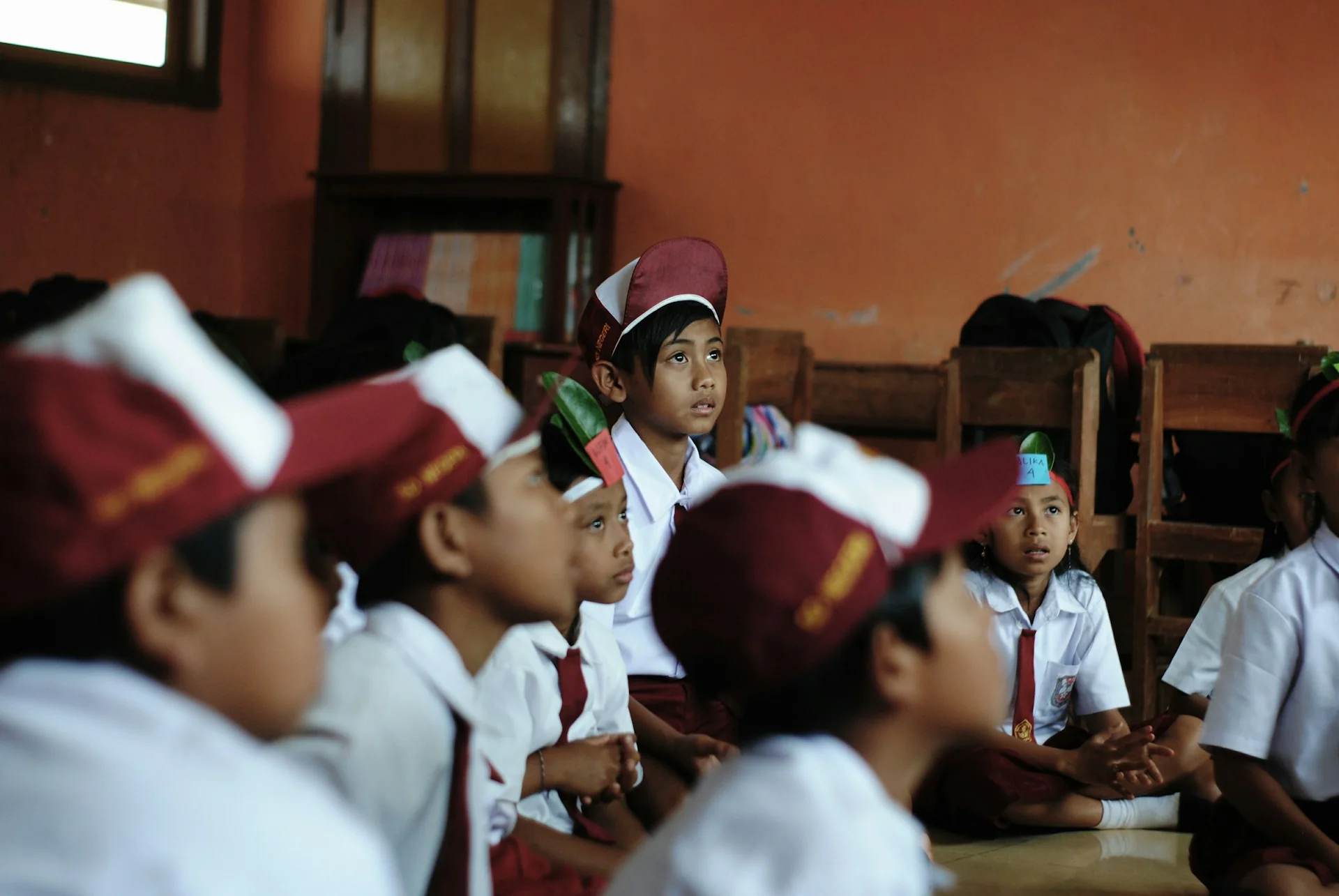 Siswa sekolah dasar berseragam putih dengan topi dan dasi merah duduk di lantai kelas