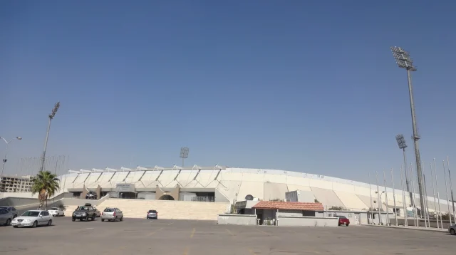 Tampak luar Stadion Internasional Amman di Yordania dengan atap tribun putih, menara lampu, dan area parkir