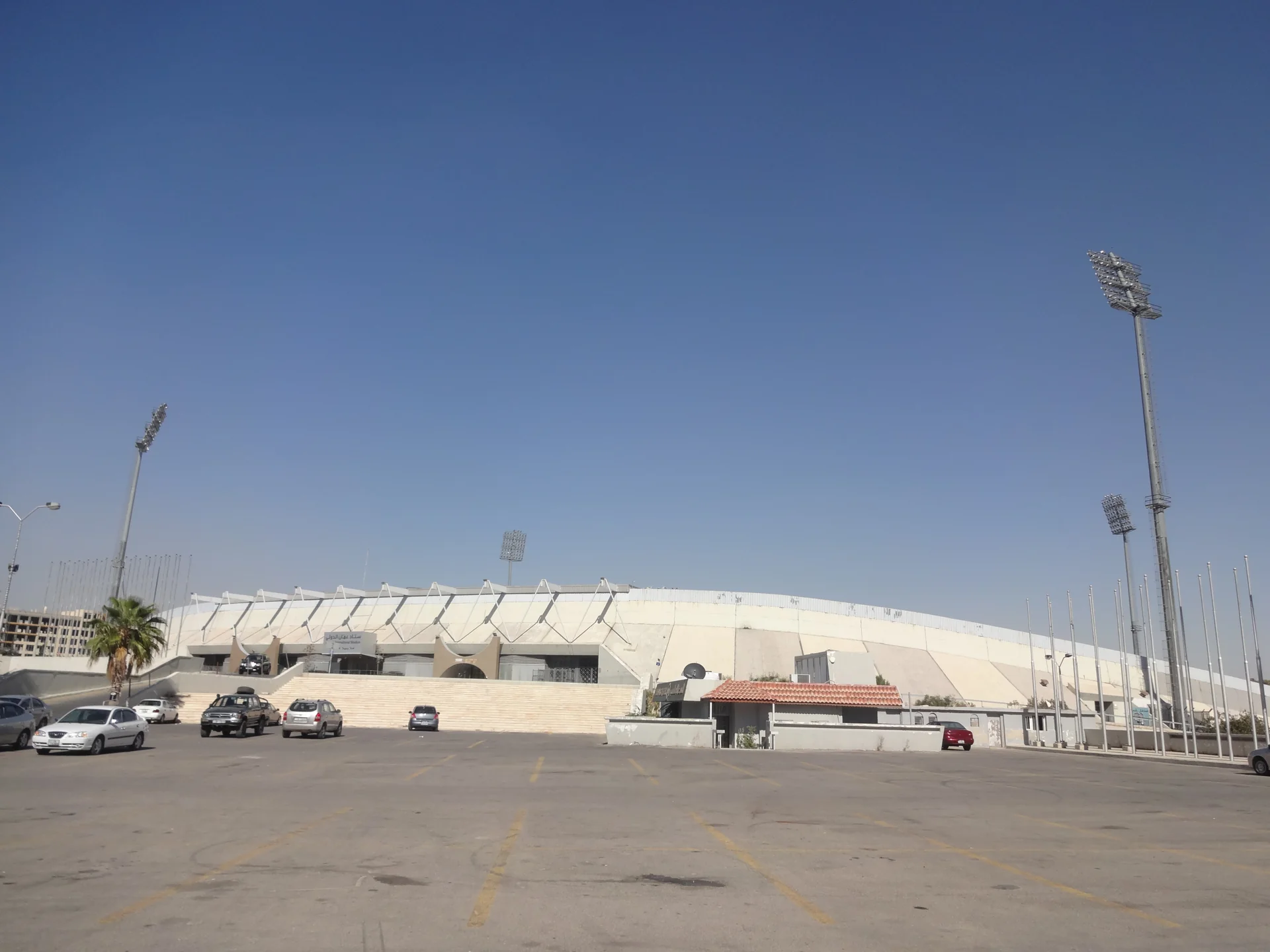 Tampak luar Stadion Internasional Amman di Yordania dengan atap tribun putih, menara lampu, dan area parkir