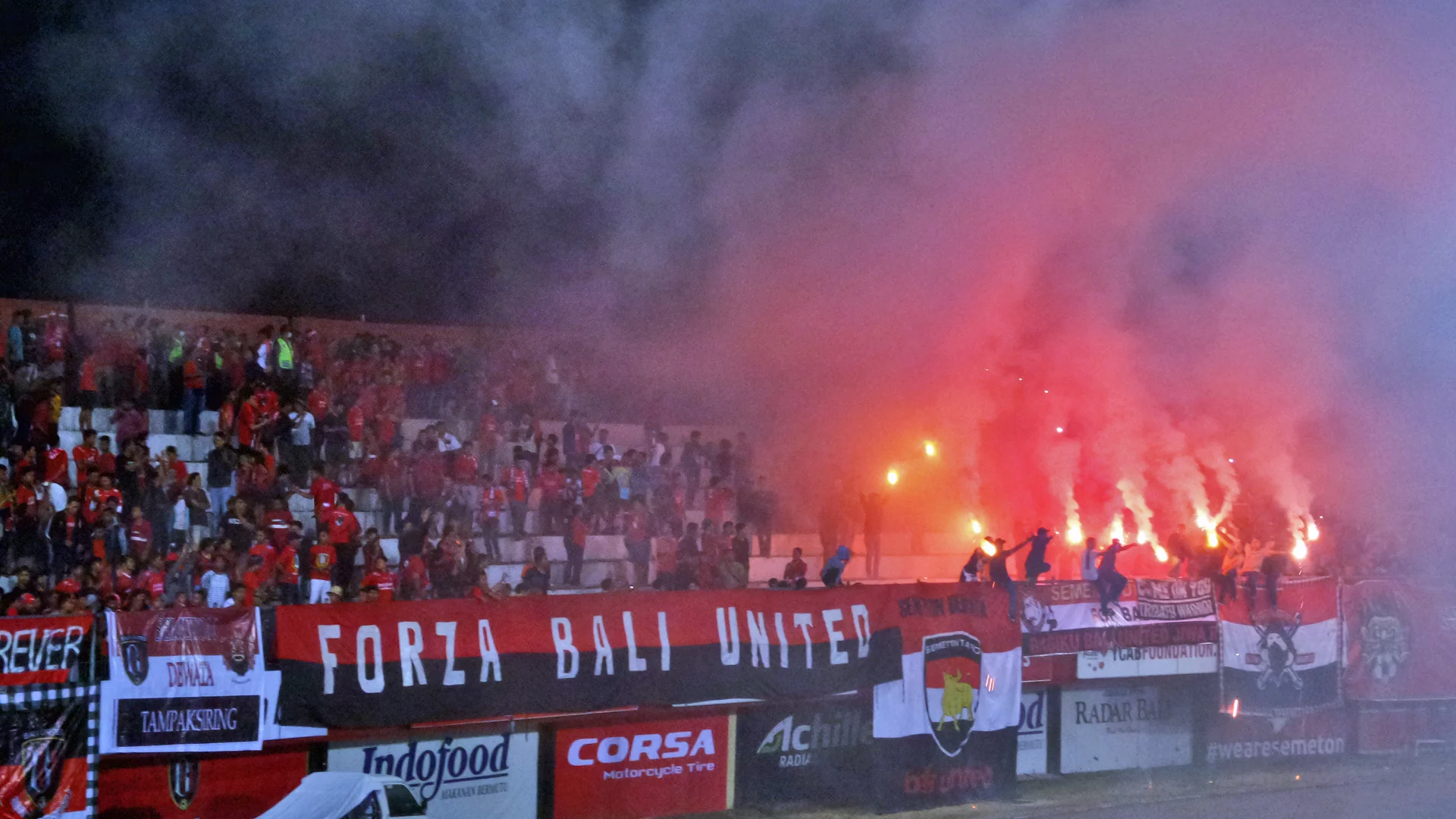 Suporter Bali United berbaju merah menyalakan suar di tribun berhias spanduk Forza Bali United