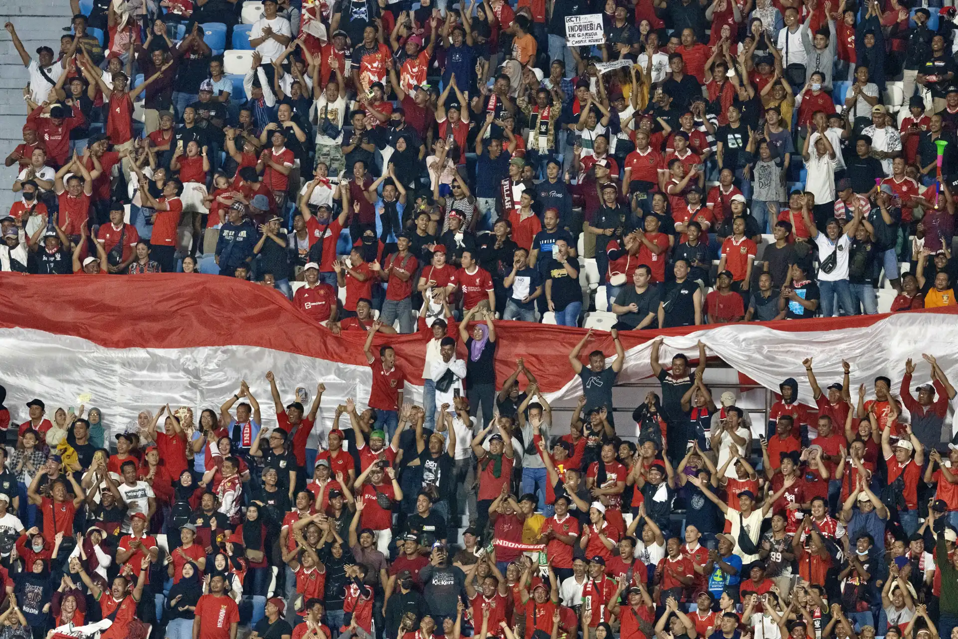 Ratusan suporter Timnas Indonesia berkaos merah mengangkat tangan di tribun sambil membentangkan bendera Merah Putih raksasa