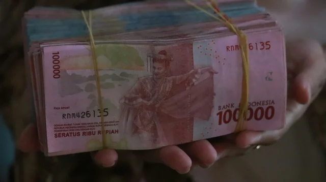 Tangan memegang gepokan uang kertas rupiah pecahan 100 ribu yang diikat karet