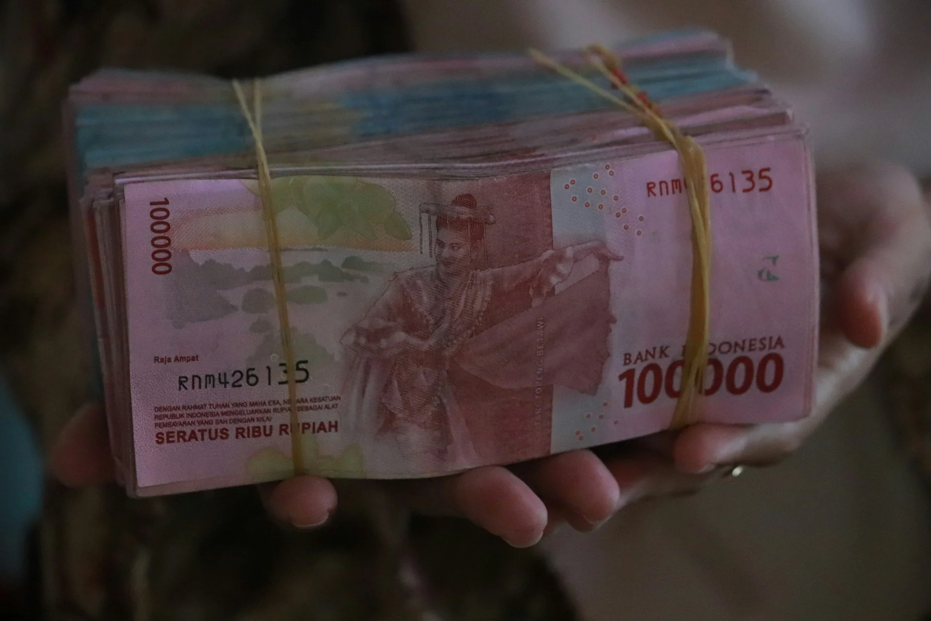Tangan memegang gepokan uang kertas rupiah pecahan 100 ribu yang diikat karet