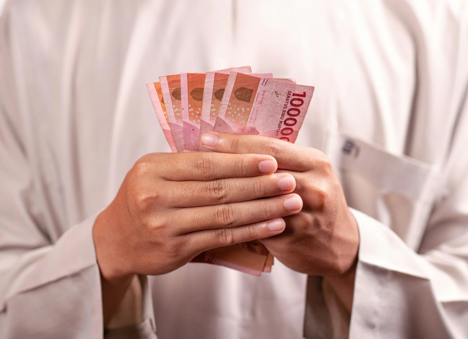 Dua tangan memegang beberapa lembar uang kertas Rp100.000 di depan kemeja putih