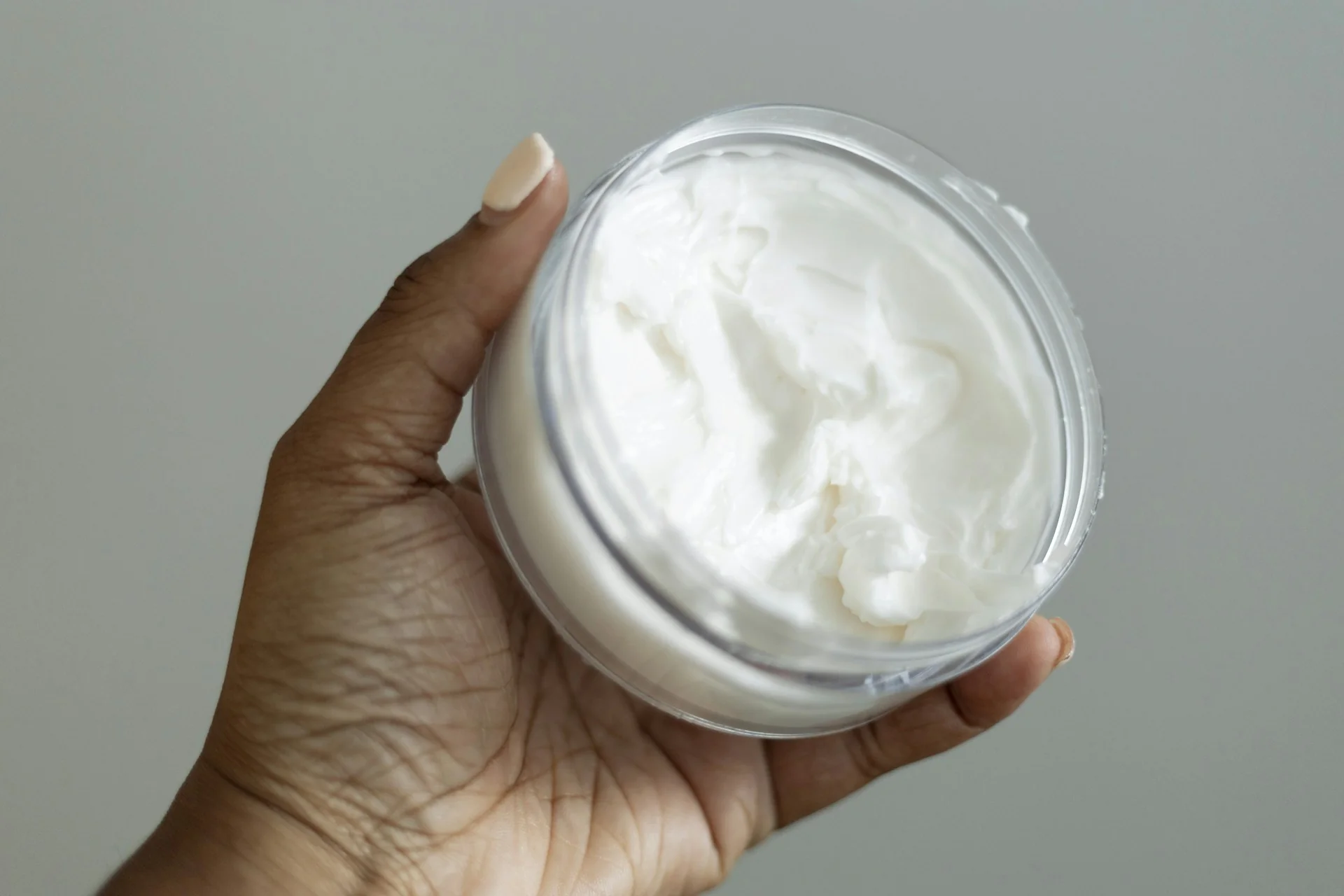 Tangan memegang wadah bening berisi krim pelembap putih bertekstur kental