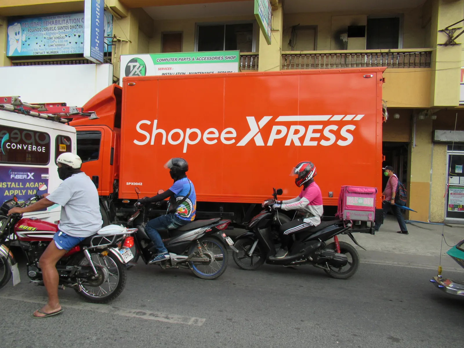 Truk oranye Shopee Xpress terparkir di tepi jalan dengan beberapa pengendara sepeda motor di depannya
