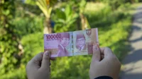 Dua tangan memegang selembar uang kertas Rp100.000 dengan latar pepohonan hijau