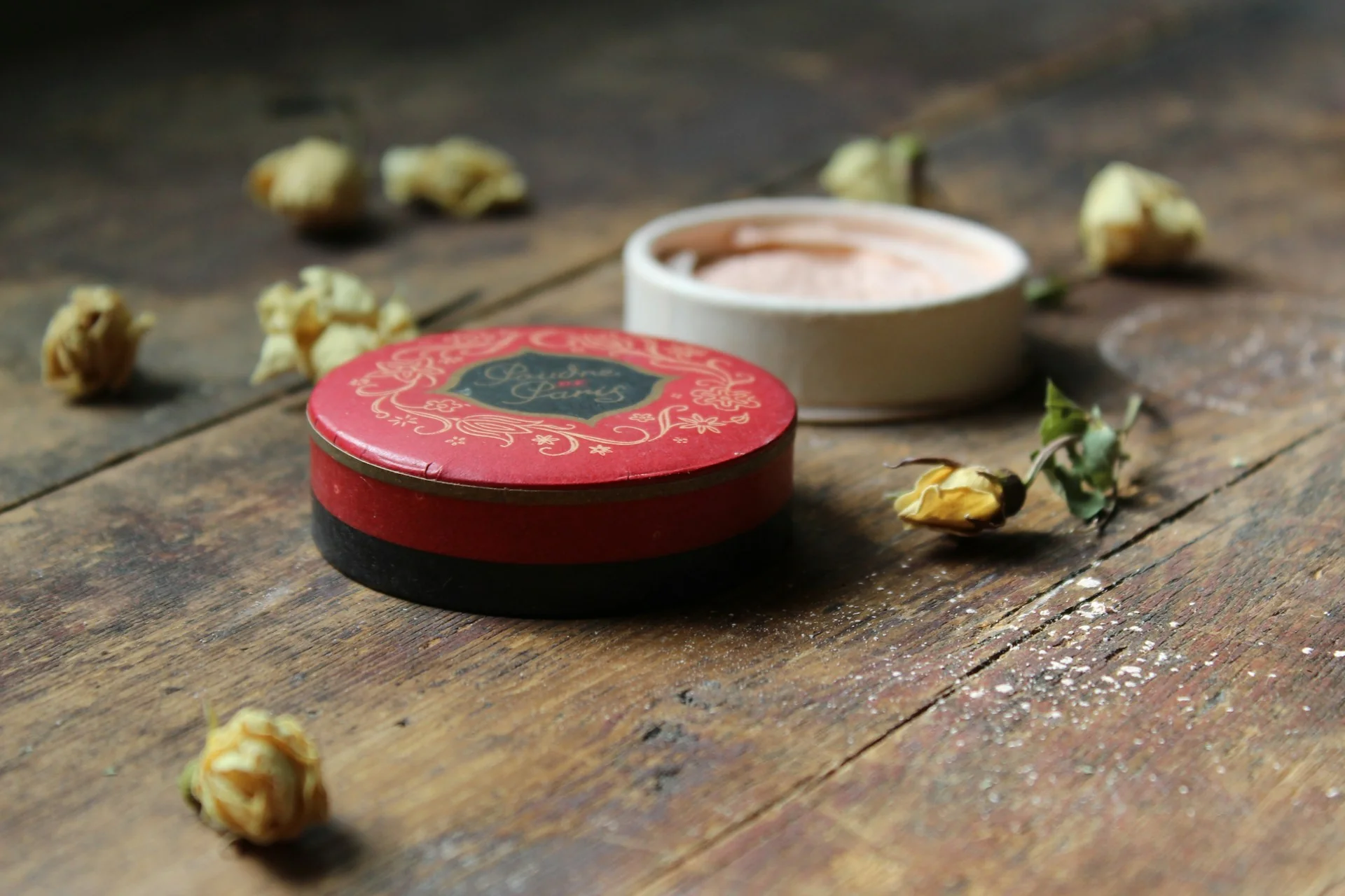 Wadah bedak bulat merah tertutup dan wadah putih terbuka berisi bedak tabur merah muda di atas meja kayu