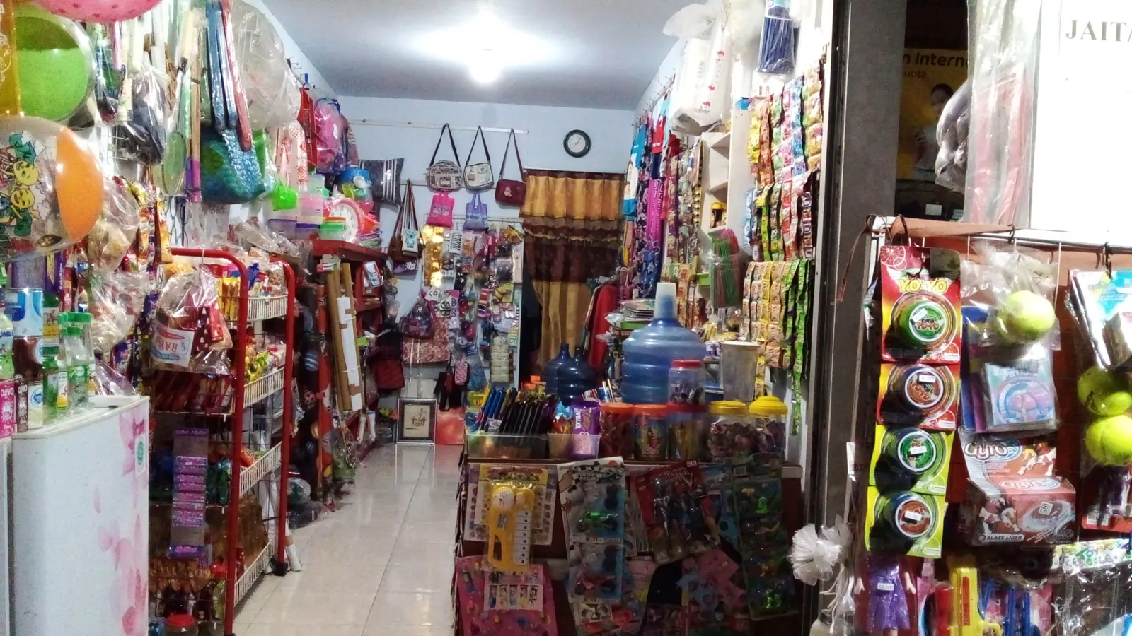 Bagian dalam warung kelontong UMKM dengan rak penuh jajanan, mainan, tas, dan galon air